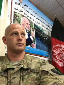 Navy officer in camouflage uniform with a poster and the Afghan flag in the background