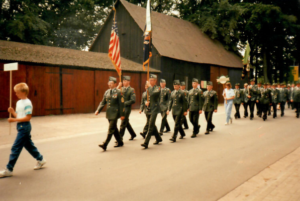 U.S. Army Soldiers in uniform carrying an American flag marching in formation down a street in a small German town.