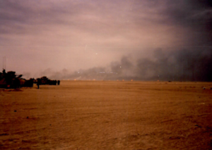 Distant oil rig fires with smoke billowing from them. Two American Army armored vehicles can be seen in the distance.