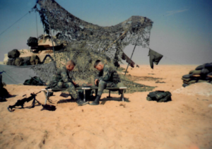 Two U.S. Army soldiers sitting on portable chairs facing each other playing a board game on a small table between them. Behind them is their Bradley Fighting Vehicle under camouflage netting.