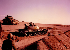 An Iraqi T55 tank in the center of the picture, with a U.S. soldier in the foreground and a Bradley Fighting vehicle behind it. The setting is the Iraqi desert.