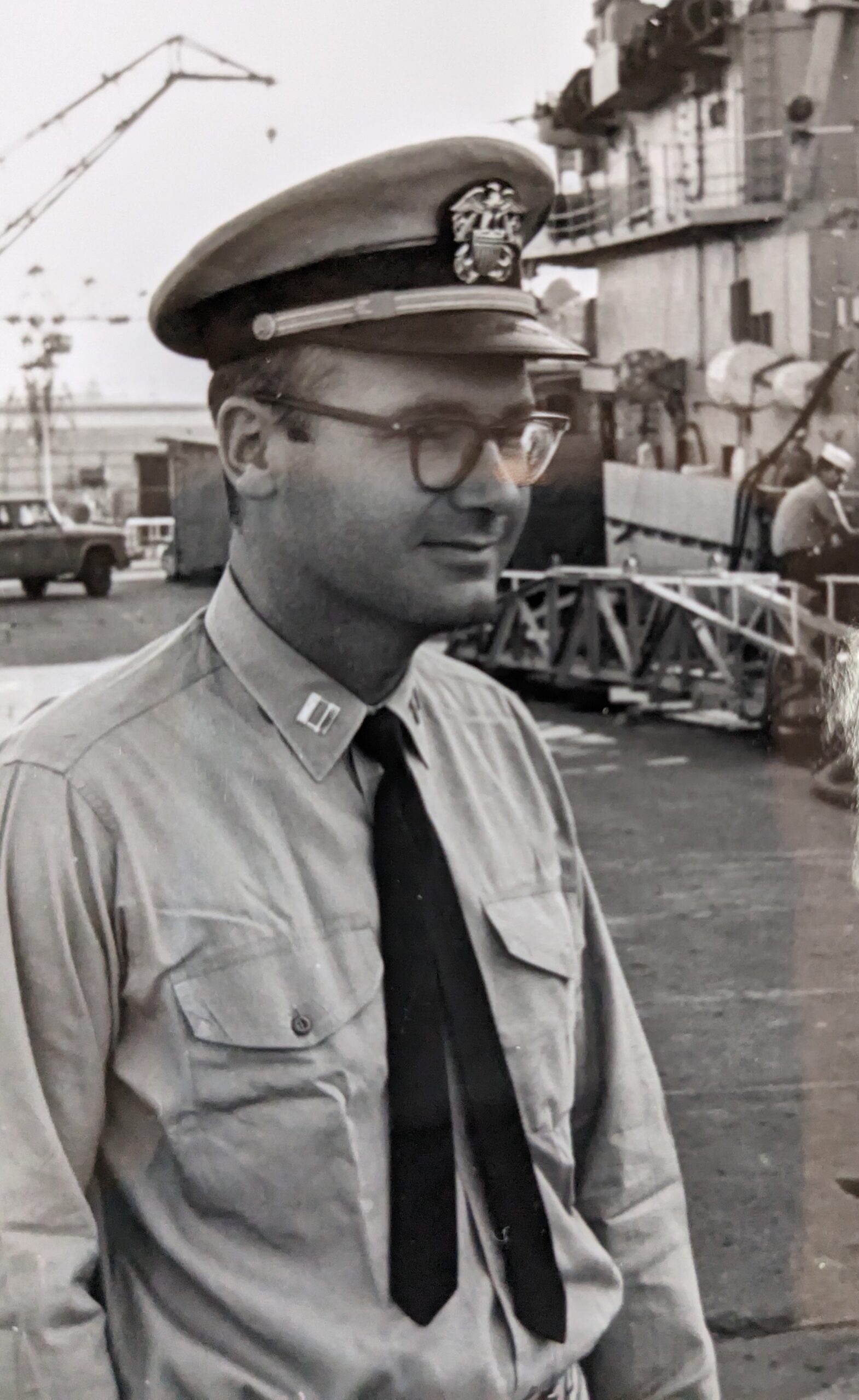 A Navy lieutenant in uniform with a ship in the background.