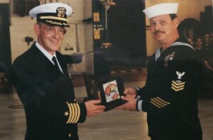 A U.S. Navy Captain presenting an award to a U.S. Navy 1st Class Petty Officer, both in dress blue uniforms