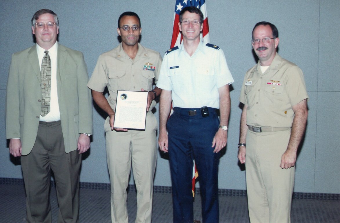 A man in a sportcoat and tie standing with two Navy officers in uniform and one Air Force officer in uniform. One of the Navy officers is holding a certificate.