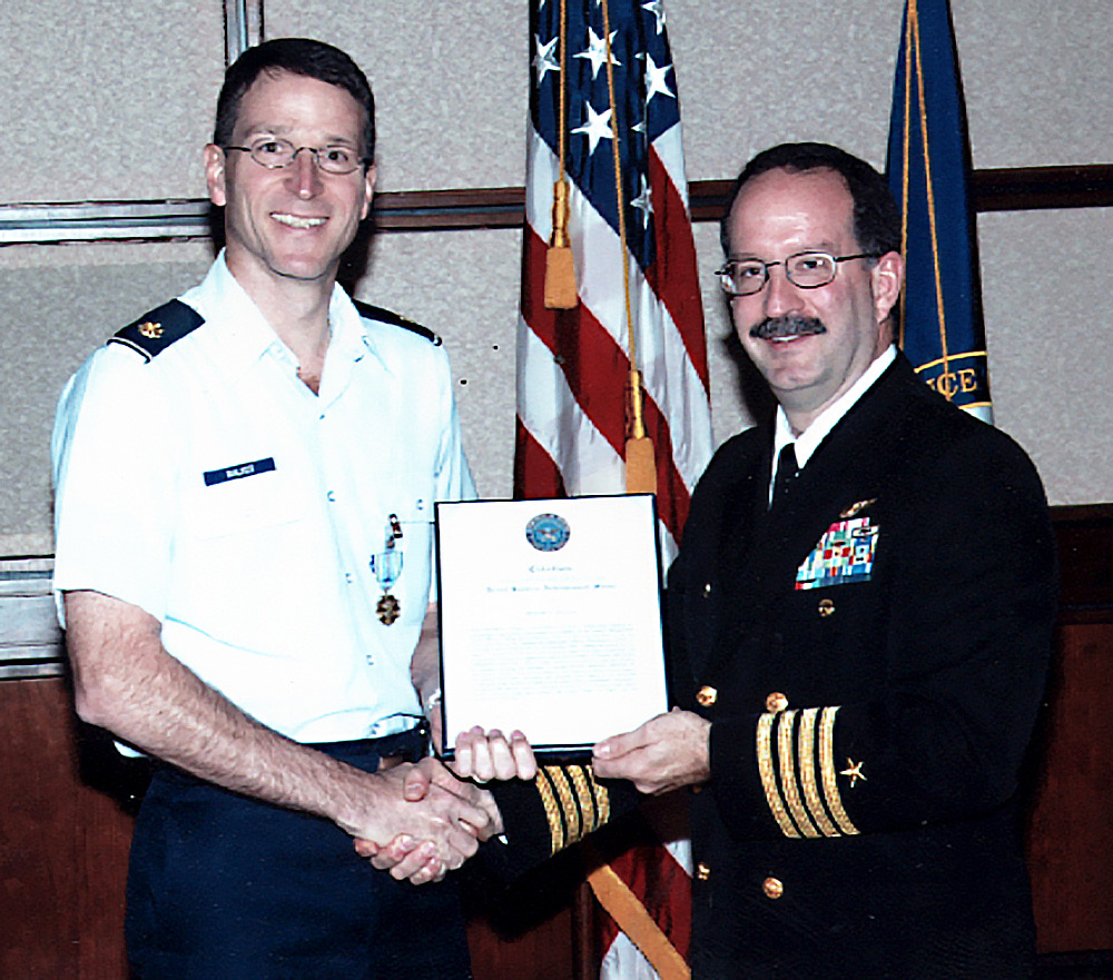 A male Air Force officer in uniform shaking hands with a male Navy officer in uniform. They are jointly holding a certificate.