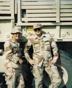Two U.S. Army soldiers in desert camouflage uniforms standing in front of a large Army truck