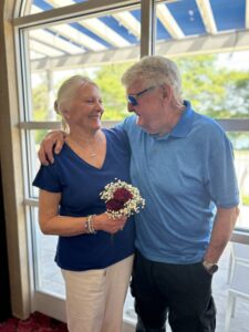 Husband with his arm around his wife. She is holding flowers and smiling and they are both looking into each other's eyes.